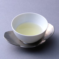 Small white porcelain cup of light color Japanese green tea on silver dual petal leaf shaped saucer on grey table