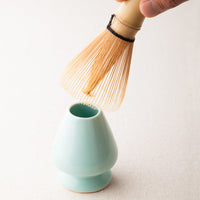 Japanese pastel blue Ippodo Tea ceramic stand and hand holding bamboo Chasen 80-tip matcha whisk nearby