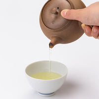 Holding lid with Ippodo Tea emblem on Yakishime Kyusu brown clay teapot and pouring light tea into white porcelain teacup