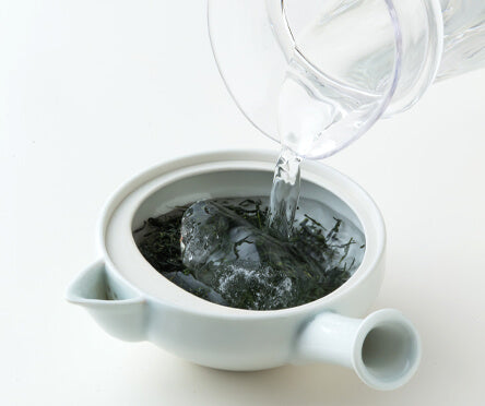 Pouring water from glass carafe into white porcelain Hasami-yaki teapot containing Gyokuro green tea leaves and ice cubes