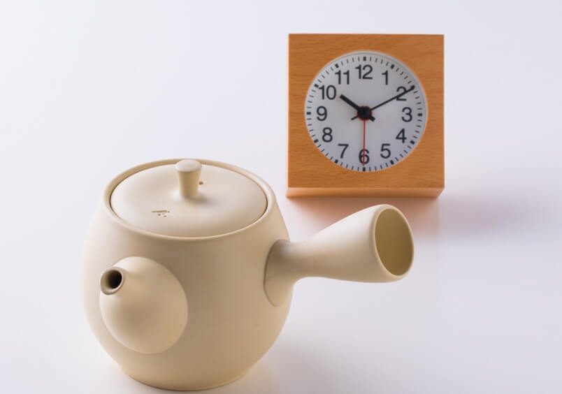 Small white porcelain Japanese kyusu teapot with blue logo on lid sitting beside orange stop watch timer on white table