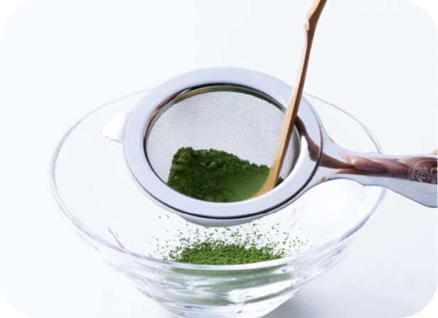 Sifting green Ippodo matcha powder through stainless steel tea strainer with bamboo tea ladel into glass bowl on white table