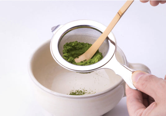 Sifting matcha through Ippodo Chakoshi tea strainer into cream ceramic teacup with handle using Chashaku bamboo tea ladle
