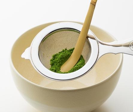 Sifting matcha through Ippodo Chakoshi tea strainer into cream ceramic teacup with handle using Chashaku bamboo tea ladle