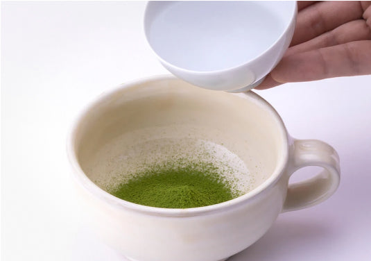 Pouring hot water from white porcelain teacup into cream colored shallow ceramic mug containing vibrant green sifted matcha