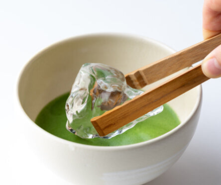 Holding clear ice cub with bamboo tongs and placing it in vibrant green matcha tea base in cream ceramic Japanese teacup