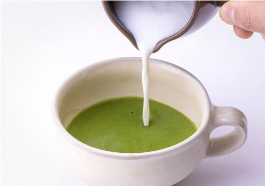 Pouring foamy frothed milk from silver pitcher into concentrated green matcha tea latte base in short beige ceramic mug