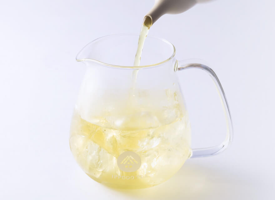 Green tea being poured from elegant spout of ivory teapot into small rounded modern glass pitcher with white Ippodo Tea logo