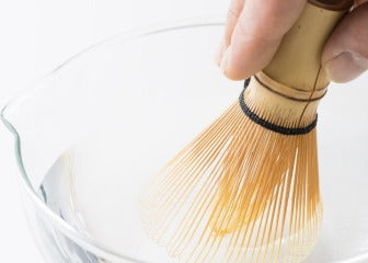 Holding tips of artisan-made Ippodo Tea Chasen 80-tip bamboo matcha tea whisk utensil in glass bowl of water with spout