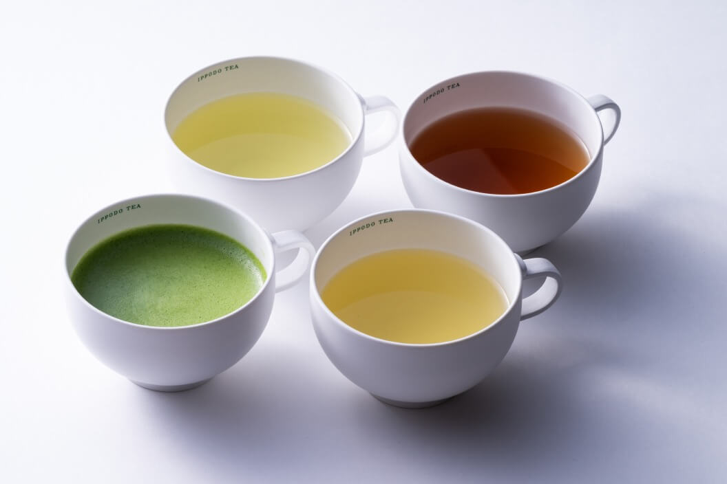 Four white teacups from Ippodo Tea containing different types of tea - one with bright green matcha, two with yellow-colored teas of slightly different shades, and one with reddish-brown tea. The cups are arranged in a square formation and photographed from above against a white background with subtle shadows.