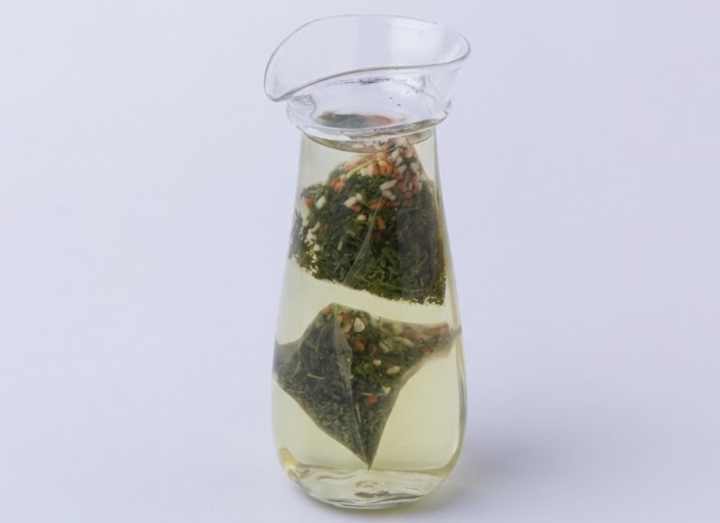 A tea bag hovering over a clear glass teapot/pitcher with a metal handle, photographed on a white background. The genmaicha tea bag appears to contain loose tea leaves and what look like small dried fruits or flowers. - Genmaicha Teabags - Cold Brew