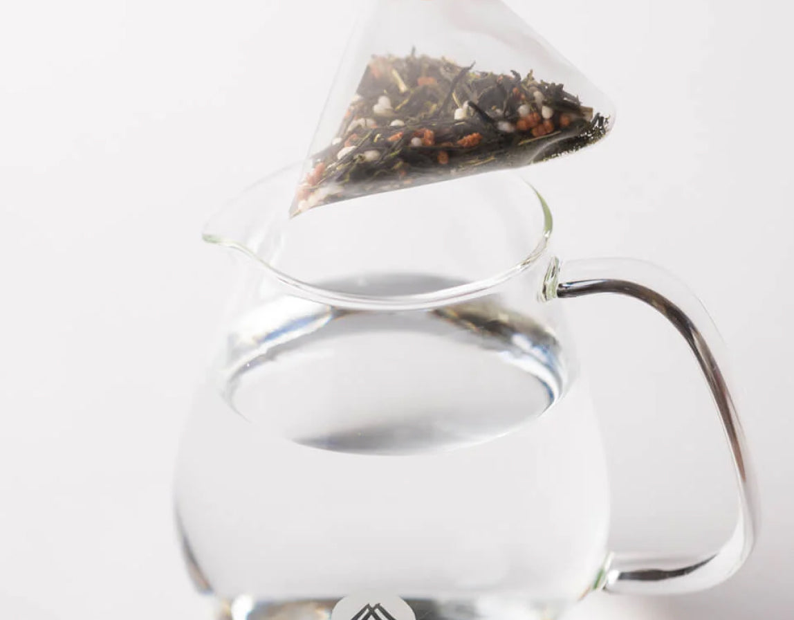 A tea bag hovering over a clear glass teapot/pitcher with a metal handle, photographed on a white background. The genmaicha tea bag appears to contain loose tea leaves and what look like small dried fruits or flowers.