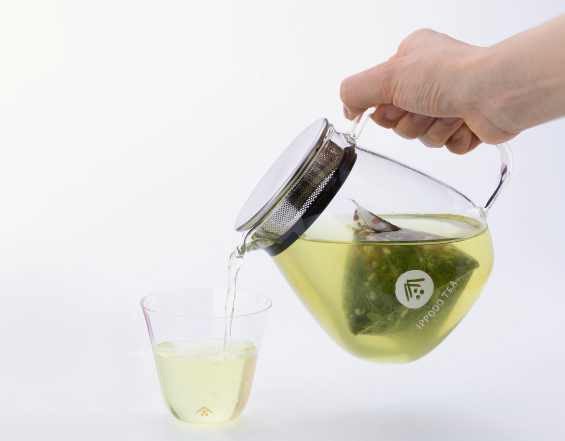 A hand pouring pale yellow-green tea from a glass teapot into a small glass cup. The teapot has a metal lid and handle, and contains what appears to be a pyramid tea bag. There's a logo visible on the teapot that reads 