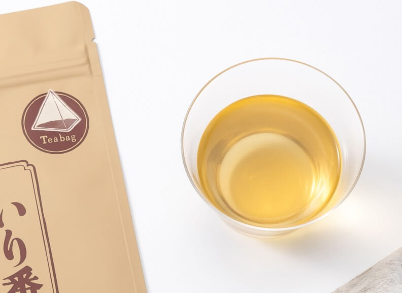 A clear glass cup filled with golden tea sits next to a tan tea bag package featuring a triangular logo. The image showcases the tea's warm color and the product's packaging, presenting a simple yet appealing composition.