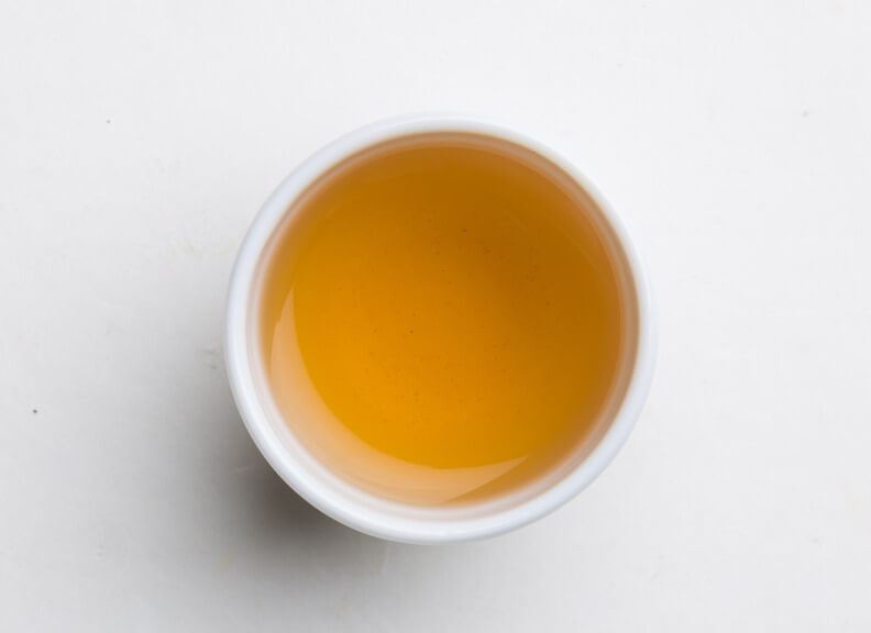 A white ceramic cup filled with golden-amber tea, viewed from above against a plain white background. The liquid's rich color contrasts with the cup's simple design, creating a minimalist and appealing composition.