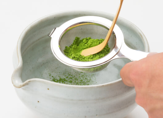 Sifting matcha through Ippodo Chakoshi tea strainer into off-white ceramic grooved tea bowl with spout using bamboo tea ladle