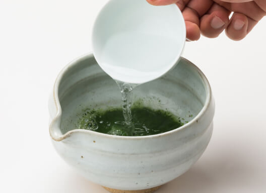 Pouring hot water from white porcelain teacup into ceramic white tea bowl with spout containing vibrant green sifted matcha