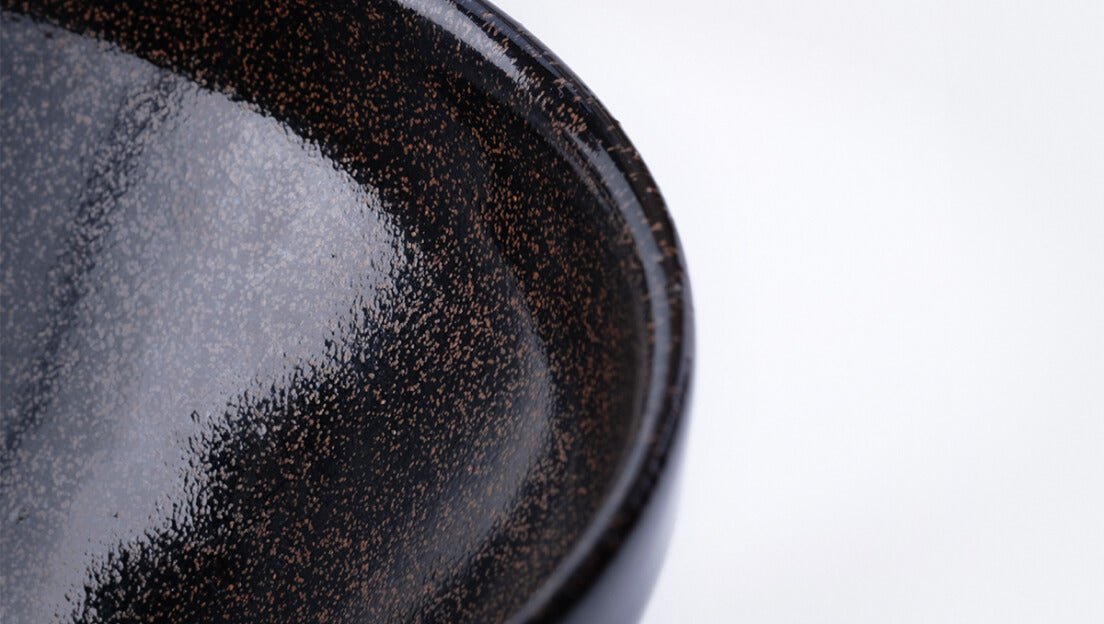 Sleek shiny cone shaped speckled black glazed ceramic matcha tea bowl with light-colored unglazed base