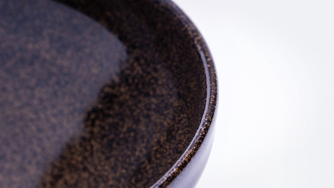 Sleek shiny cone shaped speckled black glazed ceramic matcha tea bowl with light-colored unglazed base