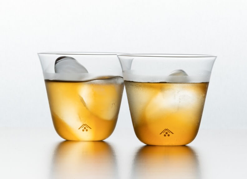 The image shows two sleek glass tumblers containing amber liquid, likely whiskey, over large round ice cubes. The glasses have a unique tapered shape and small triangular logos at the base. Set against a white background, the presentation emphasizes the elegance of premium spirits.