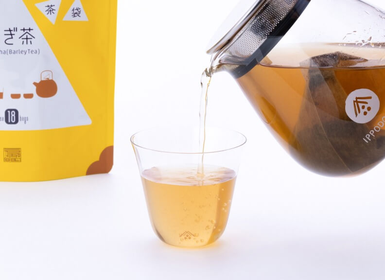 The image shows a glass teapot pouring amber-colored tea into a small, clear glass cup. A yellow tea package is visible in the background, suggesting the origin of the beverage. The modern, minimalist design of both the teapot and cup is evident, with the teapot featuring a distinctive Ippodo logo.