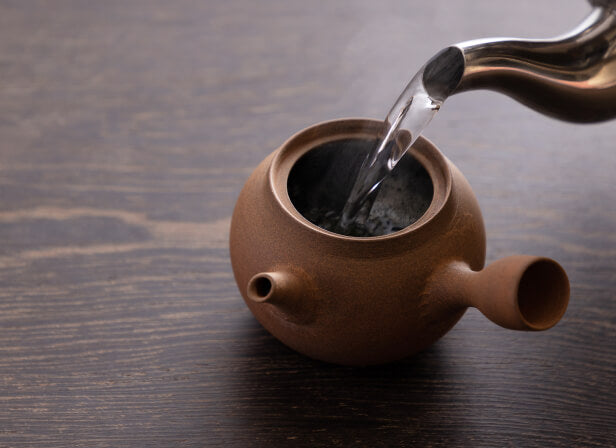 Pouring hot steamy water from stainless steel tea kettle into brown clay side-handle Japanese Yakishime Kyusu on wood table
