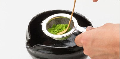 Sifting green matcha through Chakoshi tea strainer into black Ippodo Tea Bowl with Spout using Chashaku bamboo tea ladle