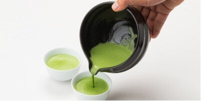 Hand pouring bright green usucha matcha from White Tea Bowl with Serving Spout into two white espresso cups on brown saucers