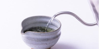 Pouring water into tea leaves in grey speckles clay tea bowl with hand groove and spout from stainless steel gooseneck kettle