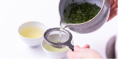 Pouring brewed looseleaf tea from grey Tea Bowl with Spout into two white porcelain teacups through Chakoshi tea strainer